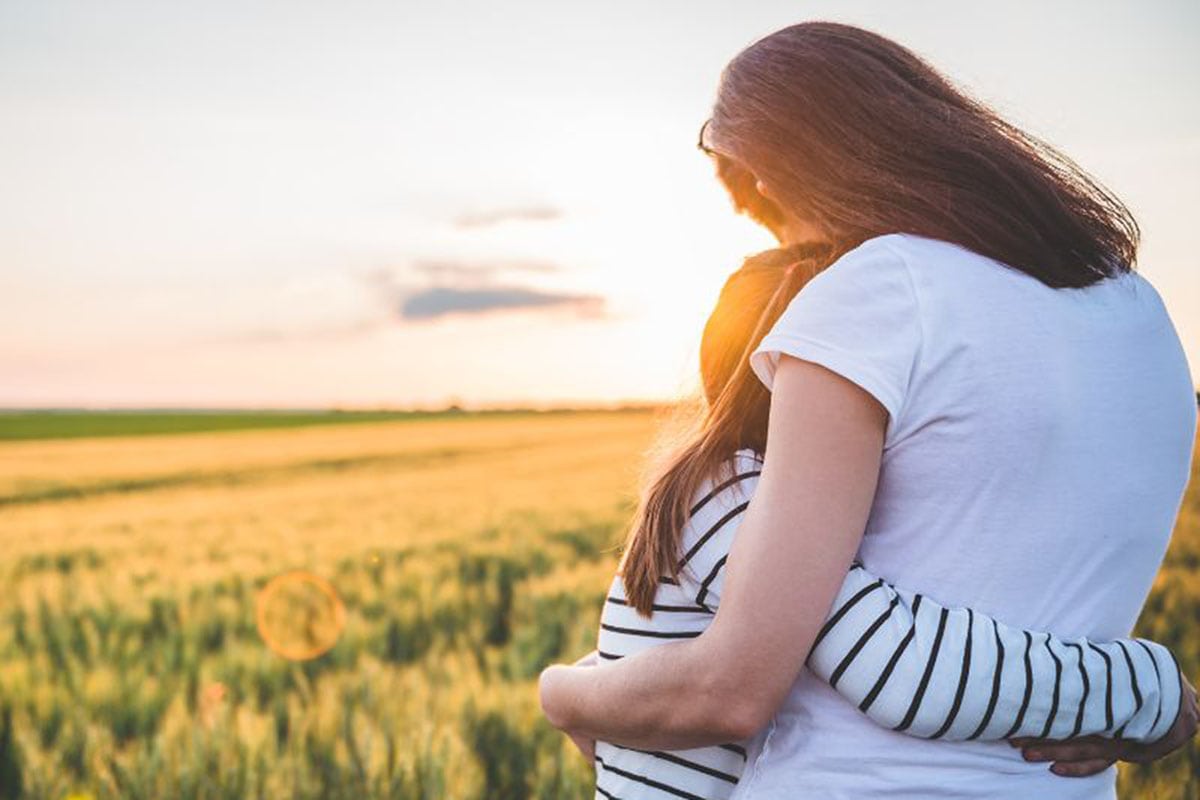 Mother hugging tween daughter in field at sunset